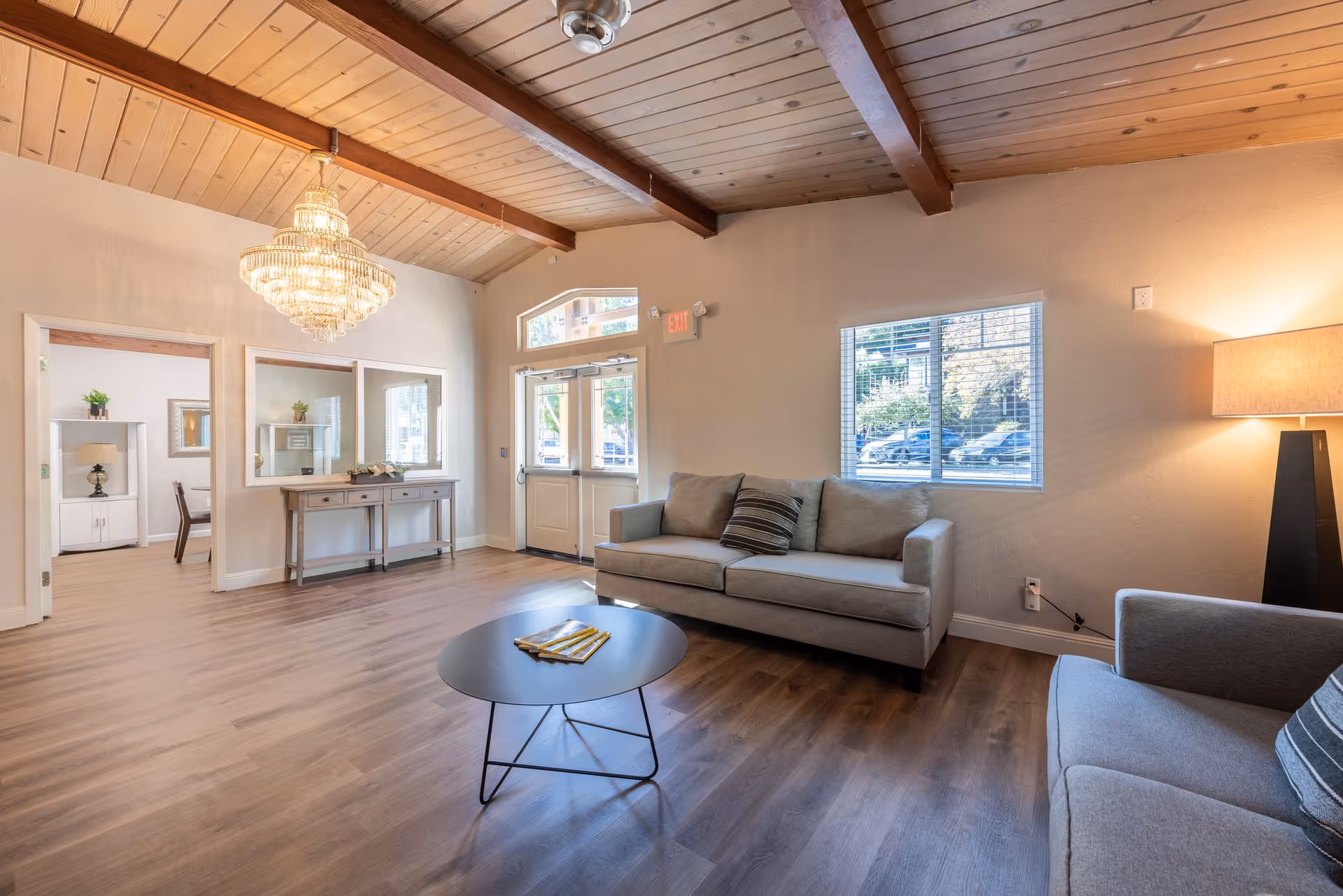 A bright and spacious living room with wooden ceiling beams, a chandelier, two gray sofas with striped pillows, a round black coffee table, a floor lamp, and large windows letting in natural light. There is a wooden console table with decorative items and a view into an adjacent room with a white cabinet and chair.