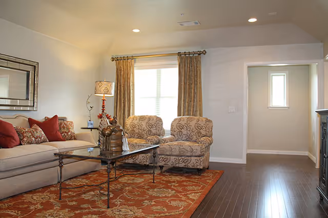 Bright living room with sofa, two patterned chairs, red rug, coffee table, and window curtains.