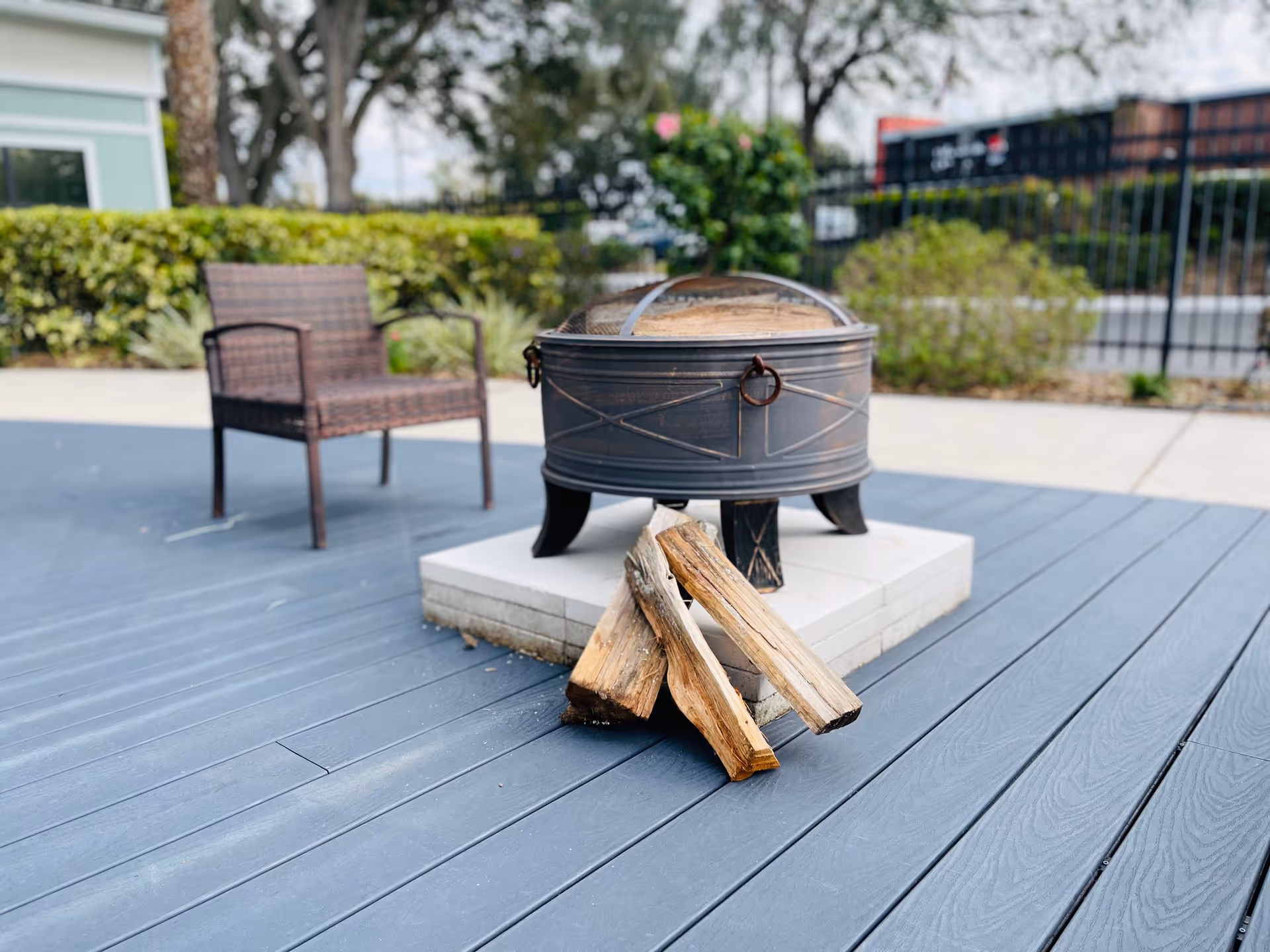 Outdoor patio area with a metal fire pit on a tiled base, three pieces of firewood leaning against it, a wicker chair nearby, and greenery and a fence in the background.