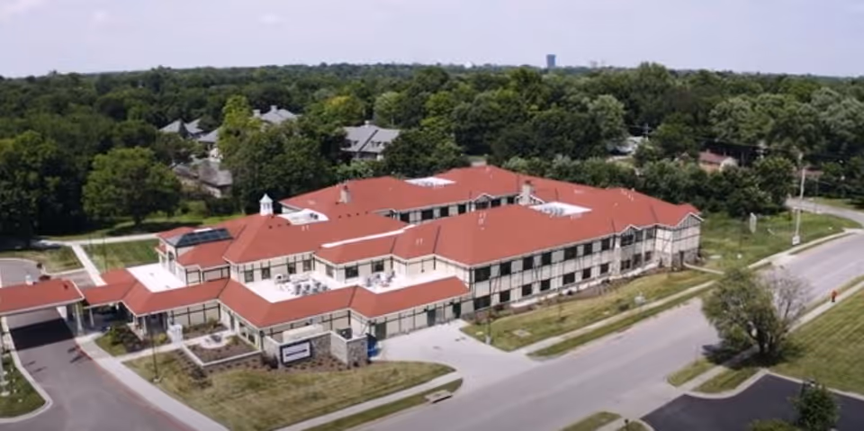 Aerial view of Creekside at Elfindale Assisted Living facility showing a large two-story building with a red roof surrounded by trees and roads.