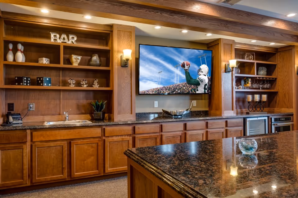 Wood-paneled communal bar area with granite countertops, built-in shelving, a sink and a wall-mounted TV showing a football game.