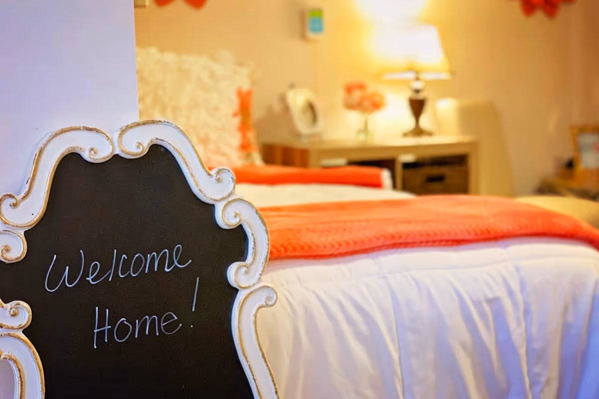 Cozy bedroom with a made bed, bedside table and lamp, and a decorative chalkboard reading "Welcome Home!"