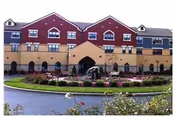 Exterior view of a multi-story residential building with a red and beige facade, featuring multiple windows and a central entrance archway. In front of the building is a circular driveway with a landscaped garden and a fountain in the center.
