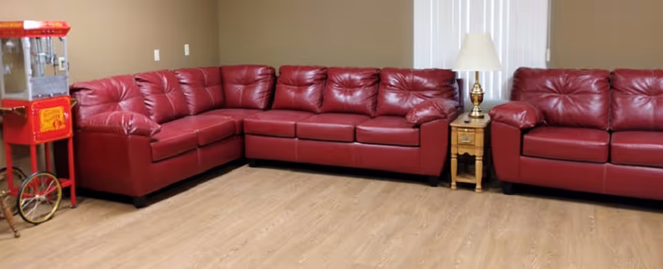 A communal lounge featuring red leather sectional sofas, a small wooden side table with a lamp, and a popcorn machine on wood flooring.