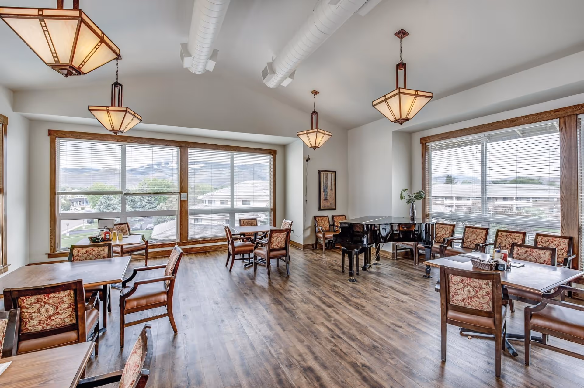 A bright and spacious dining area with wooden floors, multiple tables and chairs with patterned upholstery, large windows letting in natural light, and a black grand piano near the corner. The room features modern pendant lights and a vase with greenery on one table.