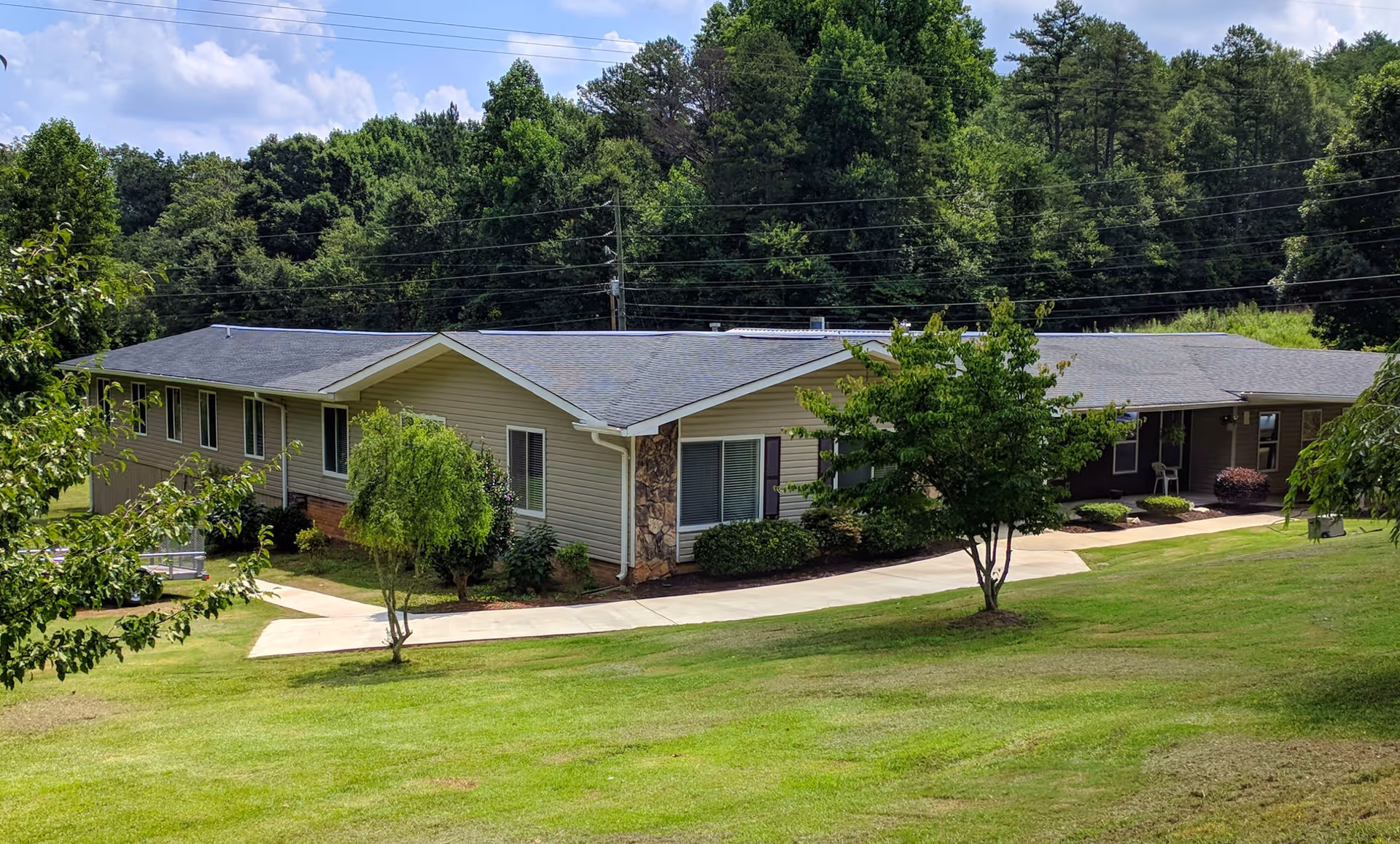 Single-story building with beige siding and a gray roof surrounded by green grass, trees, and shrubbery under a partly cloudy sky.