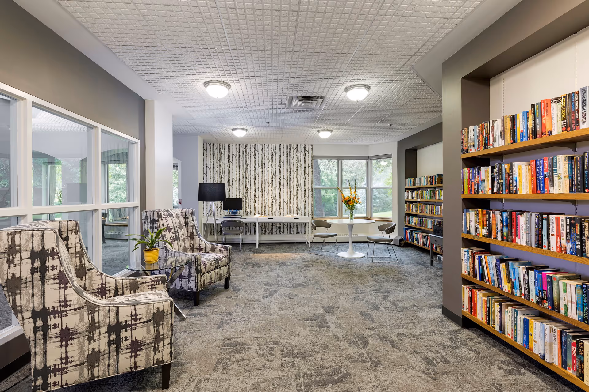 A bright and spacious reading and sitting area in a senior living facility featuring patterned armchairs, a small table with a potted plant, bookshelves filled with books, a round table with chairs near large windows, and a desk with a computer against a wall with birch tree wallpaper.