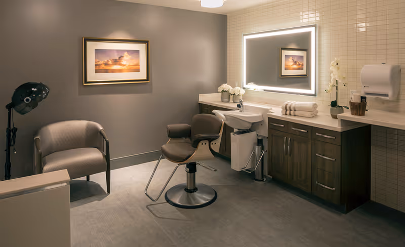 A modern salon room with a styling chair in front of a sink and a large illuminated mirror. There is a comfortable armchair to the left, a hair dryer on a stand, and a countertop with towels, flowers, and a paper towel dispenser. The walls are painted gray and tiled in beige, and a framed sunset photograph hangs on the wall.