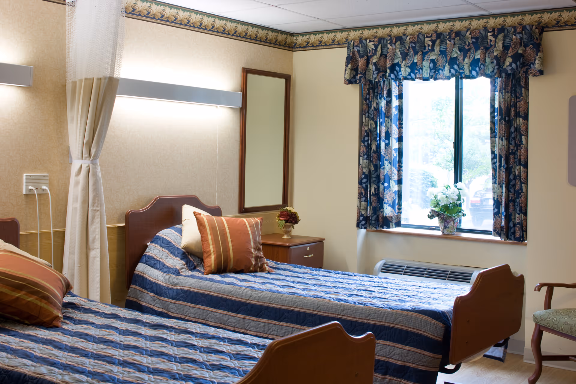 Two twin beds with blue striped bedspreads in a senior living bedroom with a window, floral curtains, and a bedside table.
