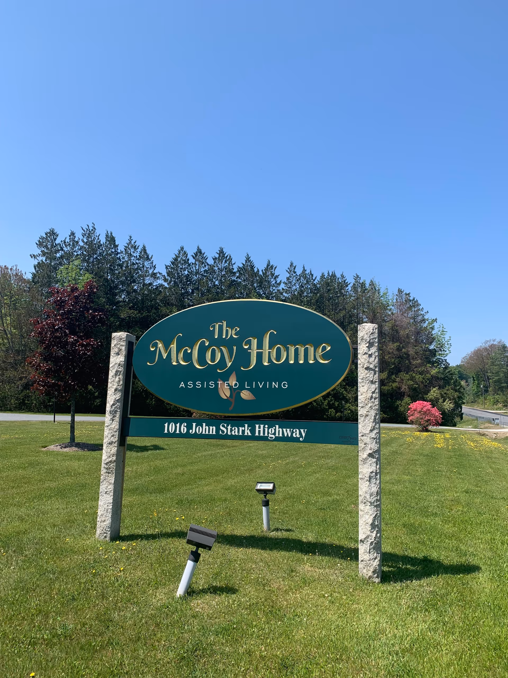 Outdoor view of a green sign for The McCoy Home Assisted Living located at 1016 John Stark Highway, set on a grassy area with trees and a clear blue sky in the background.