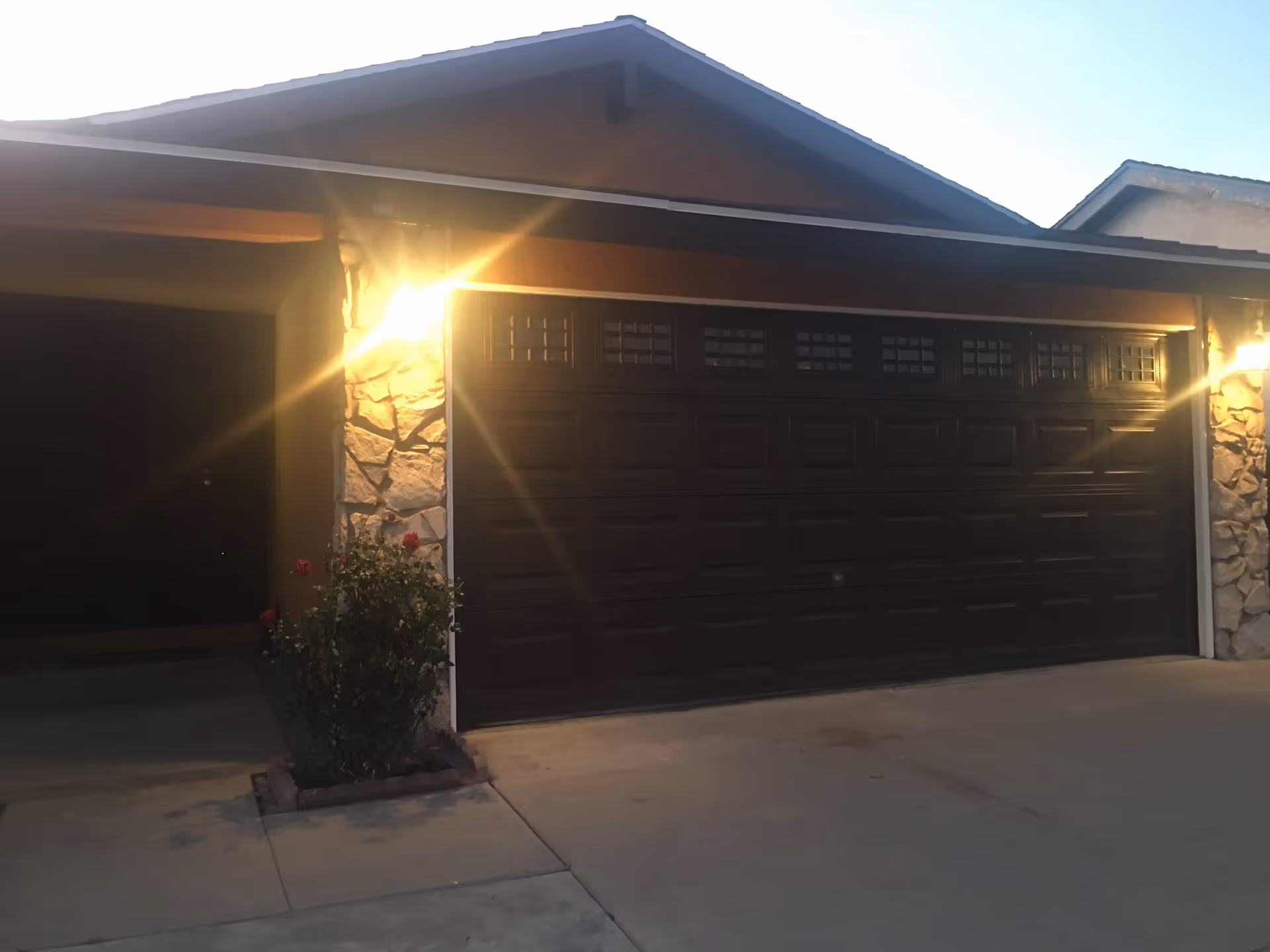Exterior view of a residential garage with a closed dark brown garage door, stone pillars on each side, two lit wall-mounted lantern lights, and a small garden bed with a rose bush in front of the garage.