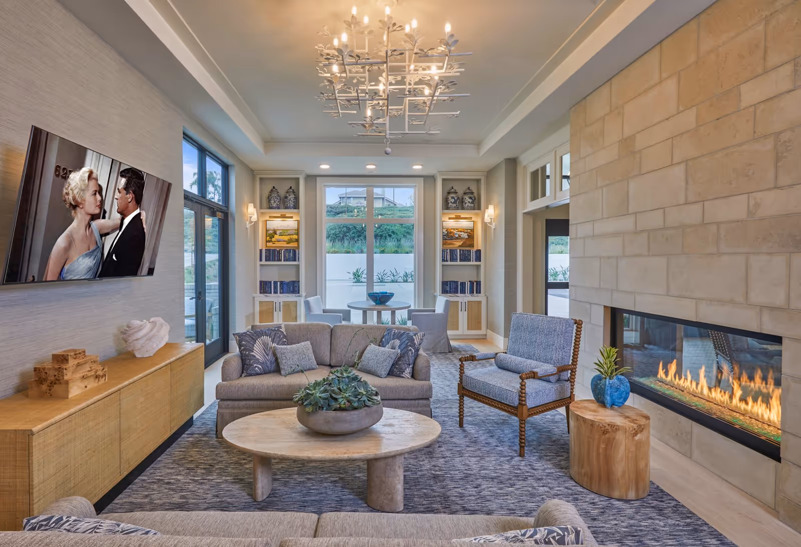 A cozy living room with a modern fireplace on the right wall, a beige sofa with patterned cushions, a wooden coffee table with a plant centerpiece, and a blue cushioned armchair with wooden frame. A flat-screen TV is mounted on the left wall showing a black and white movie scene. The room has large windows at the back with a small round table and two chairs in front of them. The ceiling features a decorative chandelier, and the room is decorated with built-in shelves holding books and decorative items.