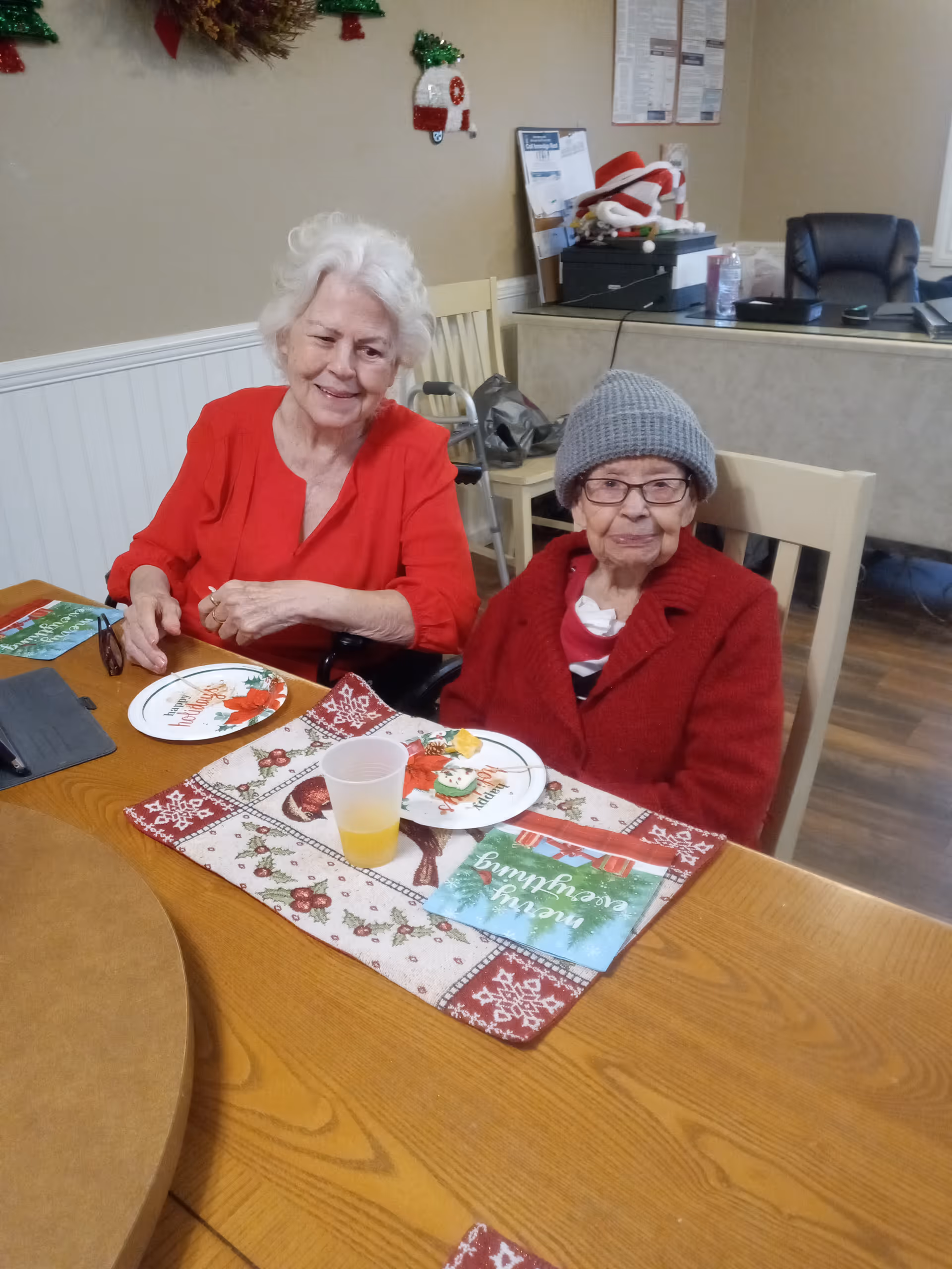 Two elderly women sitting at a wooden table in a room decorated for Christmas. One woman is wearing a red blouse and smiling, while the other is wearing a red sweater and gray knit hat with glasses. The table has holiday-themed placemats, plates, a plastic cup with a yellow drink, and a holiday greeting card. In the background, there is a desk with office supplies and holiday decorations on the wall.