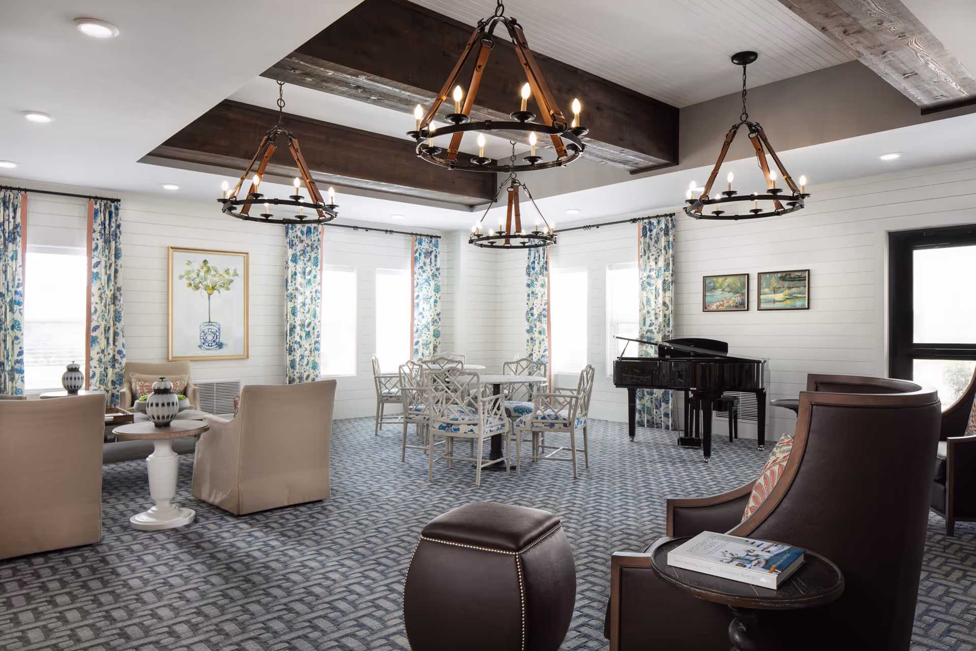 A bright and spacious living room area with patterned carpet, beige armchairs, a round table with white chairs, floral curtains on multiple windows, three large rustic chandeliers hanging from the ceiling, and a black grand piano in the corner. The walls are white with wood paneling and decorated with framed artwork.