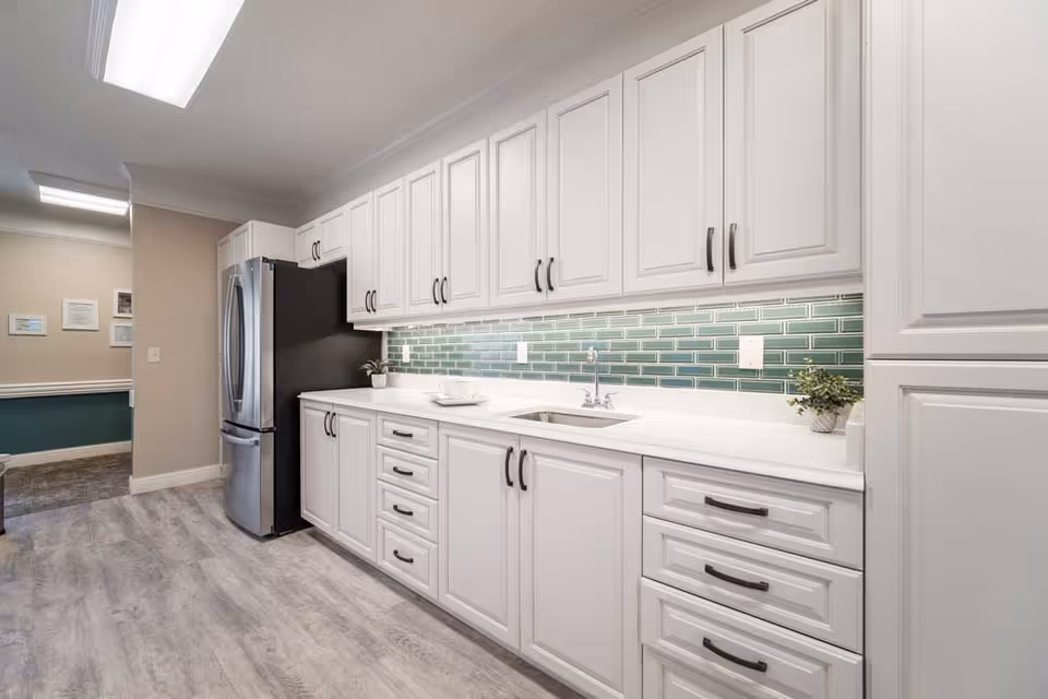A modern kitchen with white cabinetry, black handles, a stainless steel refrigerator, a white countertop with a built-in sink, and a green subway tile backsplash. The floor is light gray wood, and there is bright overhead lighting.