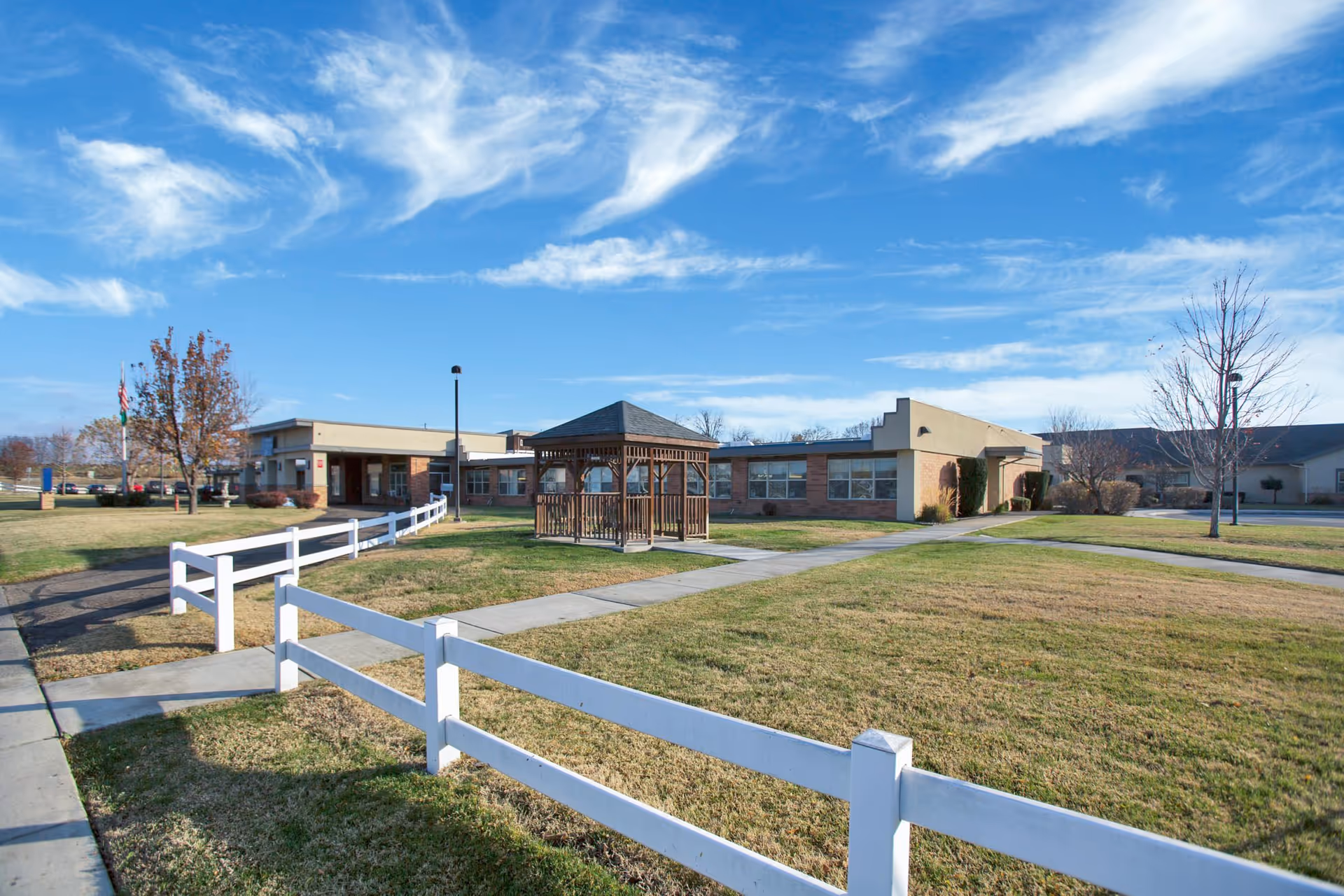 Front exterior of a healthcare facility with a lawn, white fence, and a central wooden gazebo under a bright blue sky.
