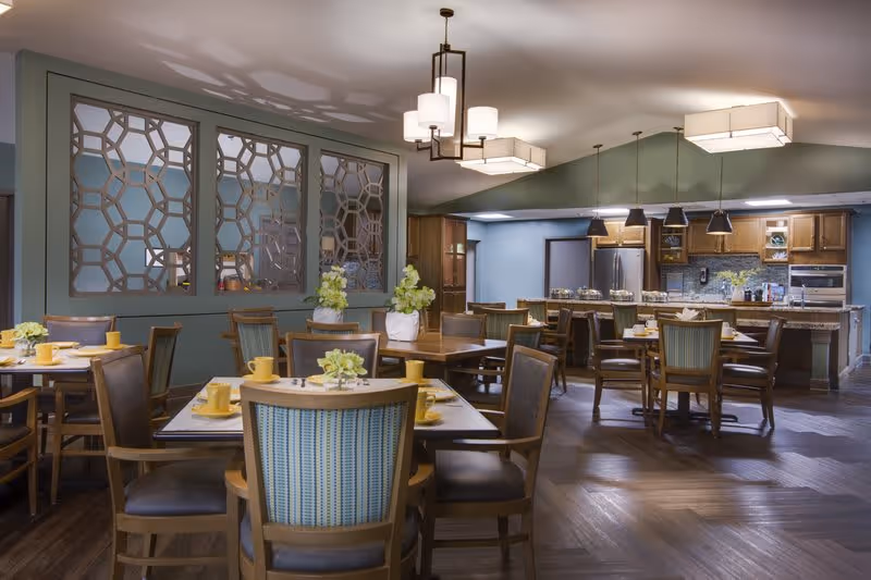 A dining area in a senior living facility with several wooden tables and chairs arranged neatly. Each table is set with yellow cups and plates, and decorated with small flower arrangements. The room features a decorative partition with geometric patterns and a kitchen area in the background with wooden cabinets, stainless steel appliances, and pendant lighting.