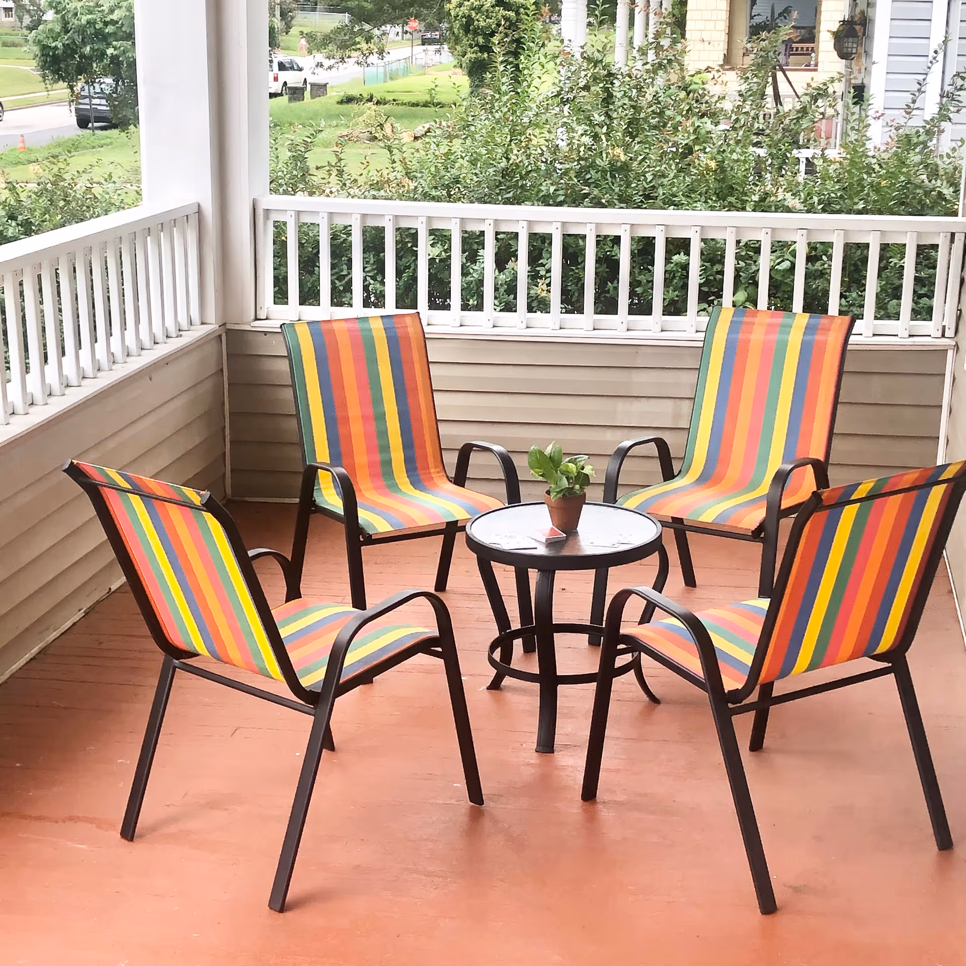 A covered outdoor porch area with four colorful striped chairs arranged around a small round glass table with a small potted plant on top. The porch has white railing and beige siding, with greenery and a street visible in the background.