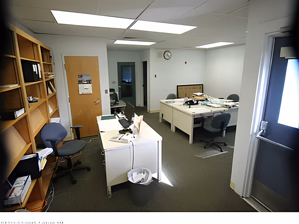 An office room with white desks, office chairs, a wooden shelving unit filled with files and papers, and a trash bin. The room has white walls, a closed wooden door, a clock on the wall, and fluorescent ceiling lights. Papers and office supplies are scattered on the desks.