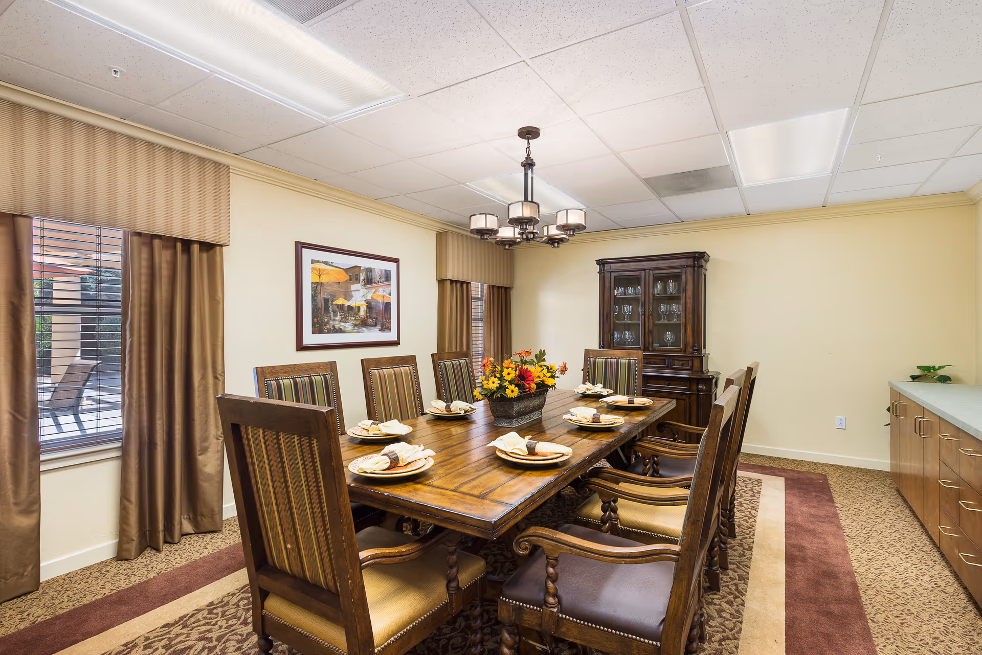 A formal dining room with a large wooden table set for eight people. The table has plates with napkins and a centerpiece of colorful flowers. The room features striped upholstered chairs, a wooden china cabinet with glassware, beige walls, carpeted floor with a patterned rug, and two windows with brown curtains. A chandelier with multiple lights hangs above the table.