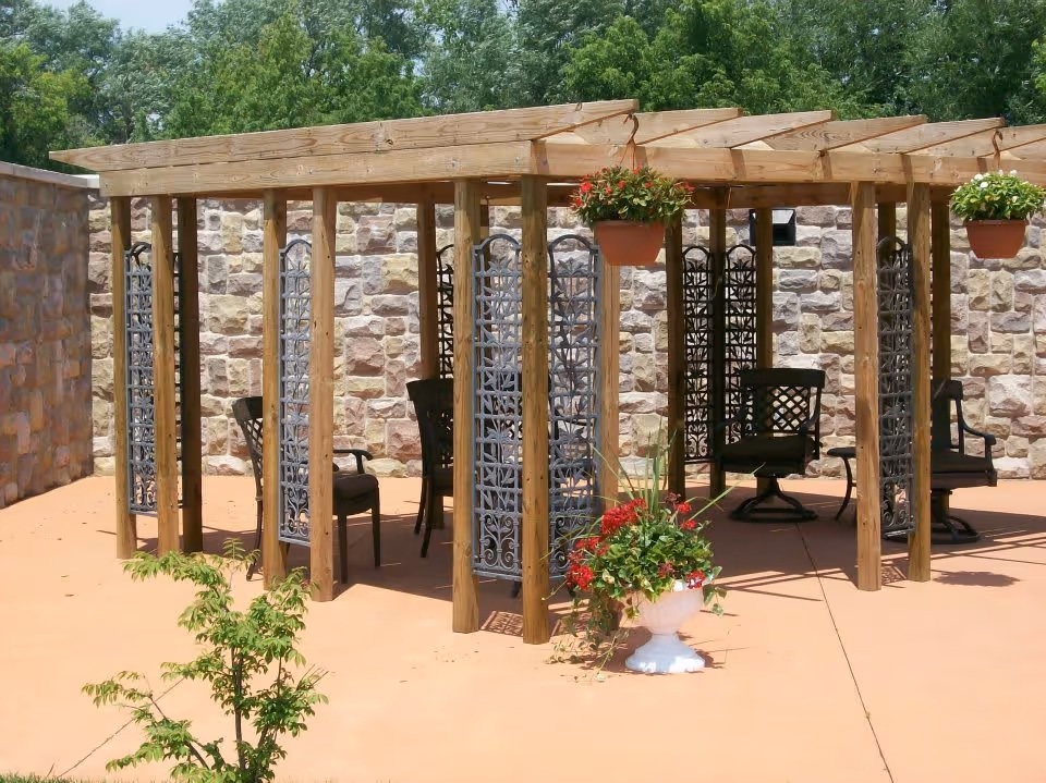 Outdoor patio area with a wooden pergola structure featuring decorative metal panels and several black chairs underneath. There are hanging flower pots with red and green plants attached to the pergola, and a white planter with red flowers and greenery on the ground. The background shows a stone wall and green trees.