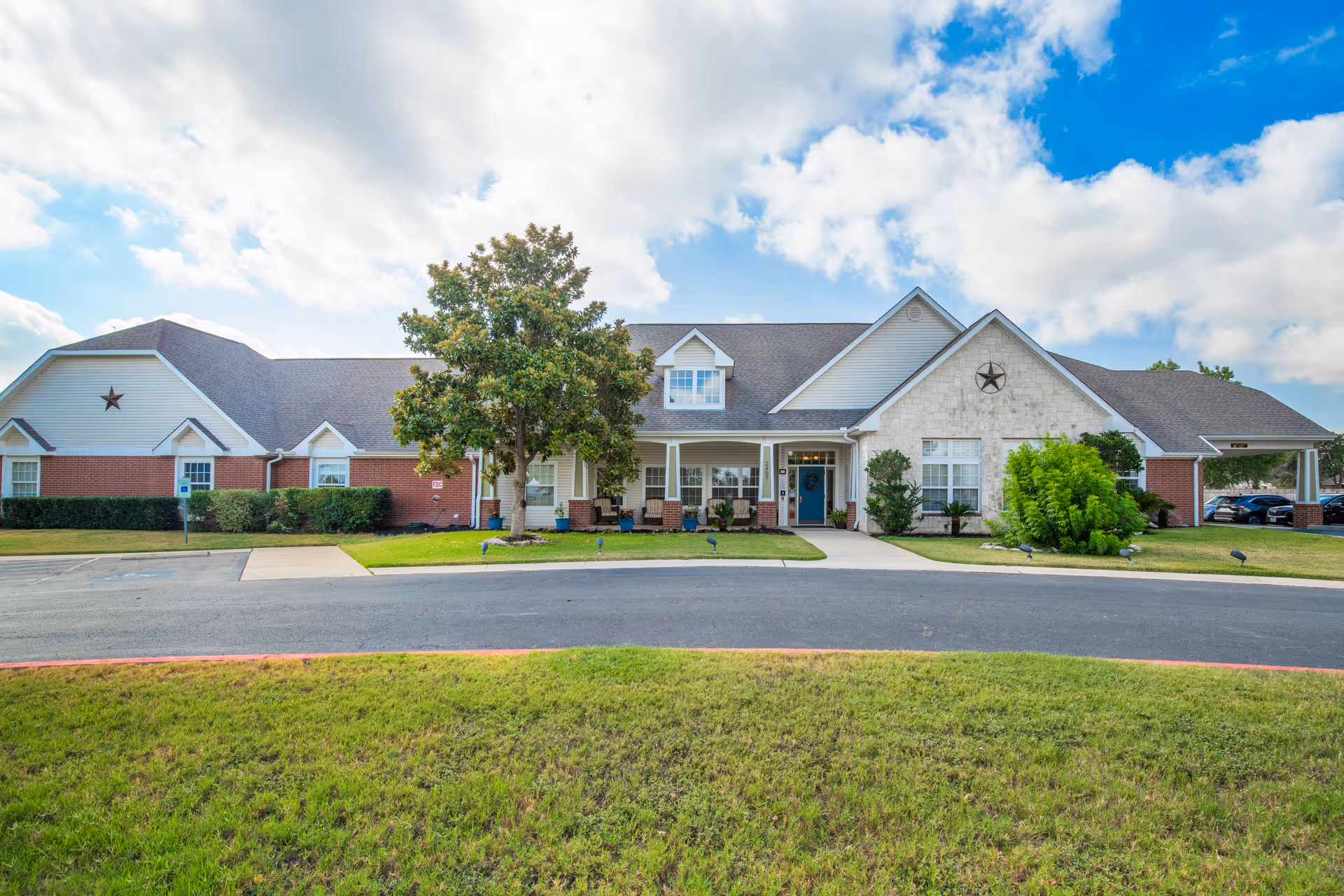 Front exterior of a single-story senior living building with a covered entrance, porch seating, and landscaped lawn under a partly cloudy sky.