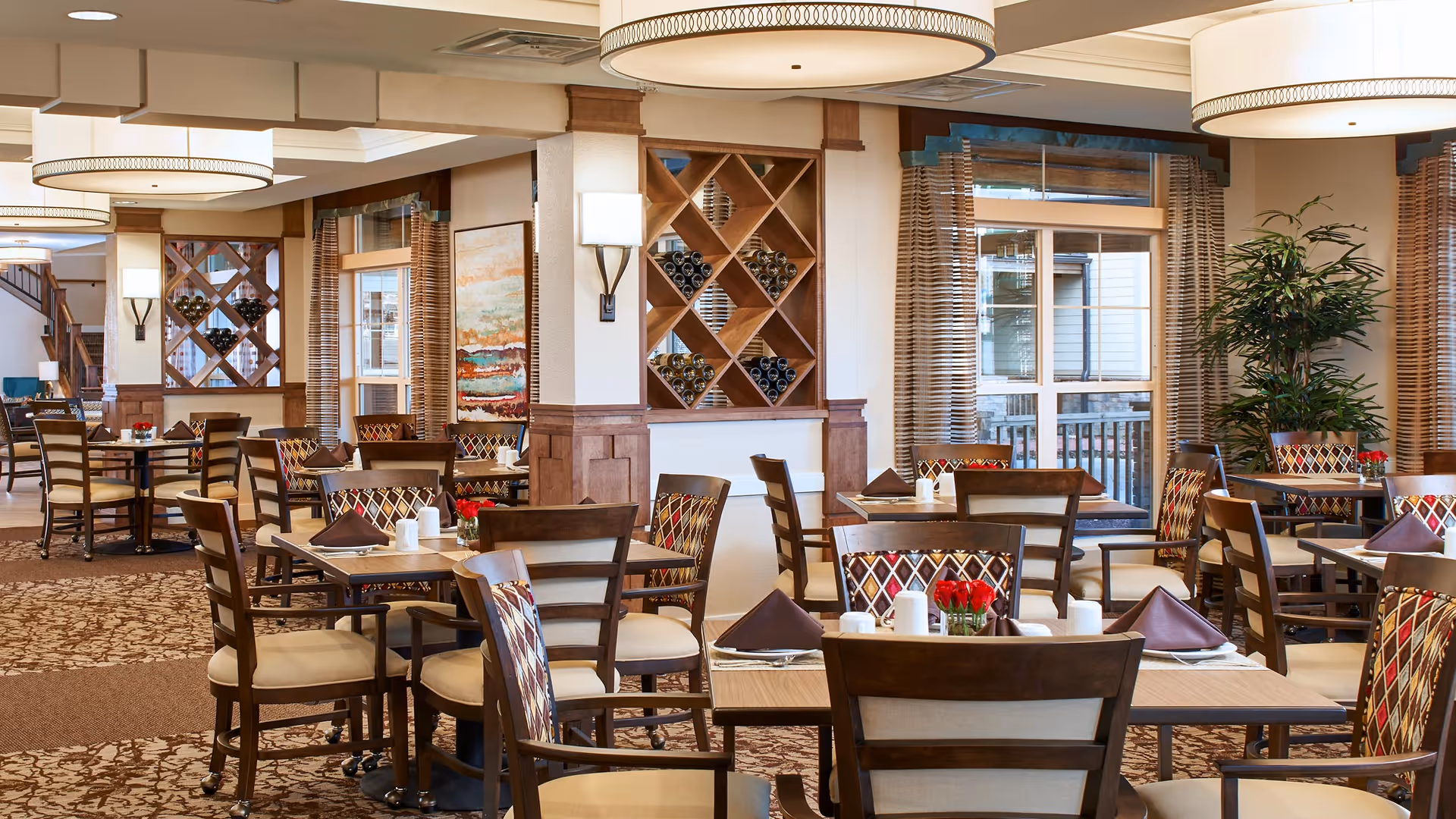 A well-lit dining room in an assisted living facility featuring multiple wooden tables and chairs with patterned upholstery. Each table is set with brown napkins, white salt and pepper shakers, and small vases of red flowers. The room has large windows with striped curtains, decorative wall sconces, and built-in wine racks. The carpet has a floral pattern, and there are large circular ceiling lights providing illumination.