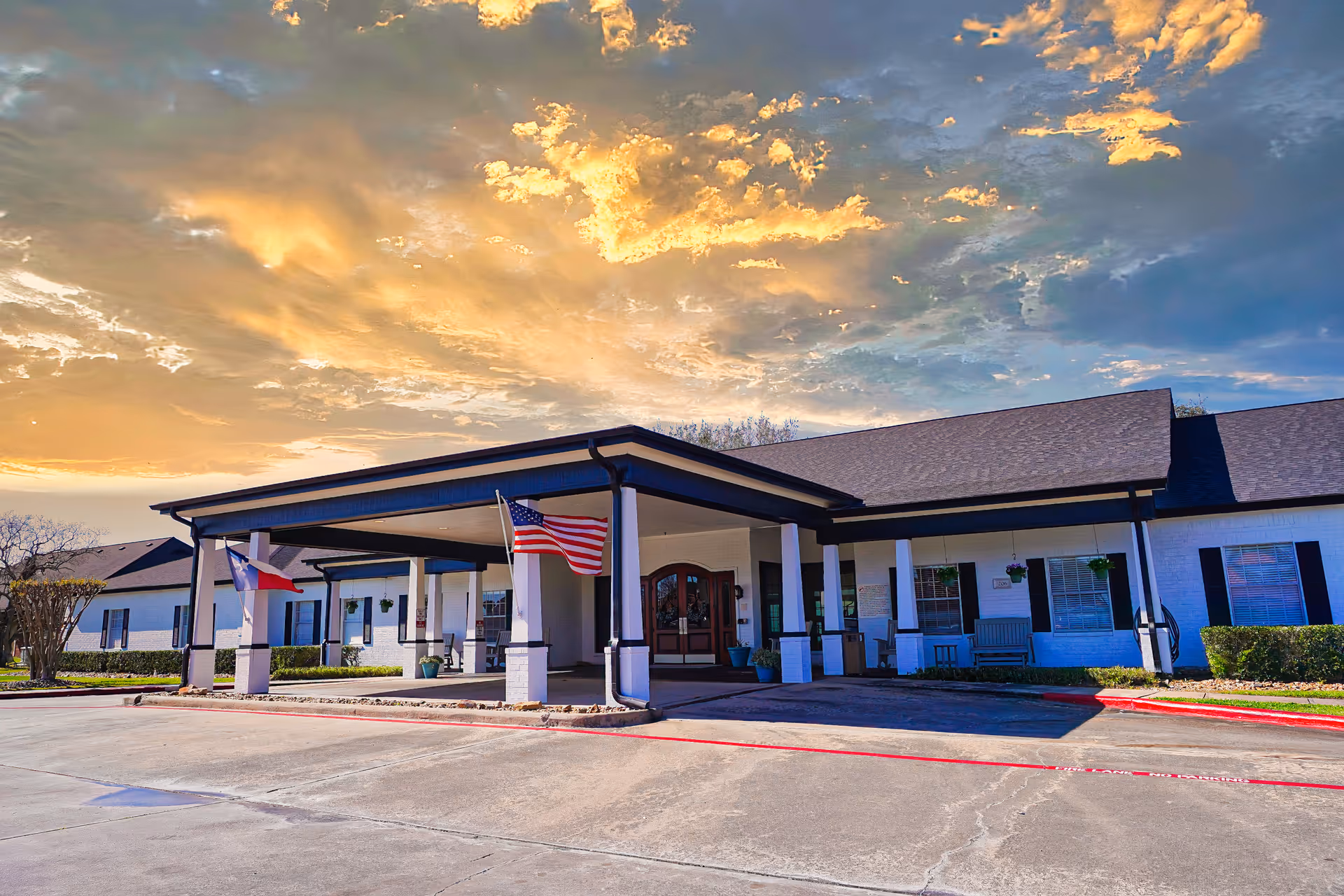 Exterior view of Sodalis Lake Jackson facility at sunset, showing a single-story building with a covered entrance supported by white columns. Two flags, the American flag and the Texas state flag, are displayed near the entrance. The sky is partly cloudy with warm golden light from the setting sun.