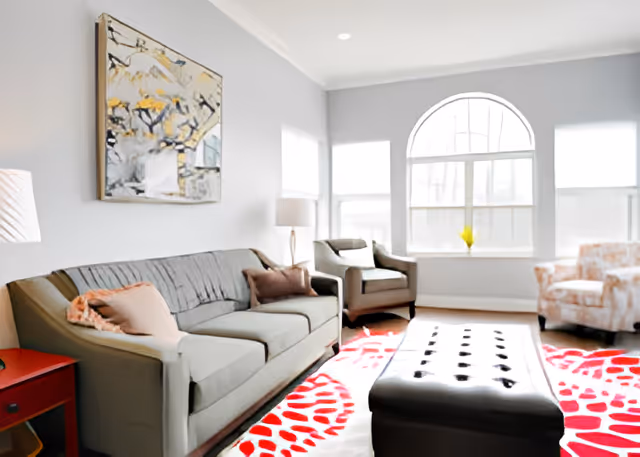 Bright living room with a sofa, armchairs, a tufted ottoman, and a large arched window over a red patterned rug.