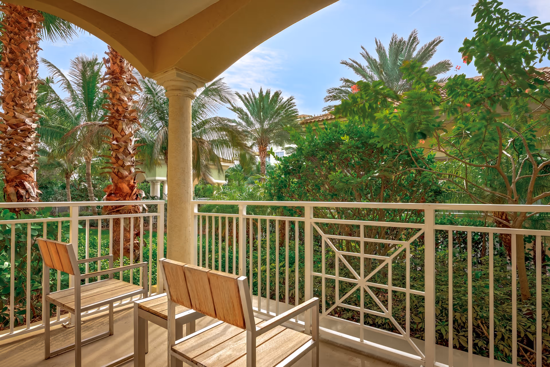 Covered balcony with two wooden chairs overlooking tropical landscaping and palm trees.