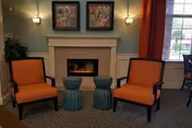 Cozy sitting area with two orange armchairs facing a lit fireplace, two small blue stools, framed artwork above the mantel, and a window to the right.