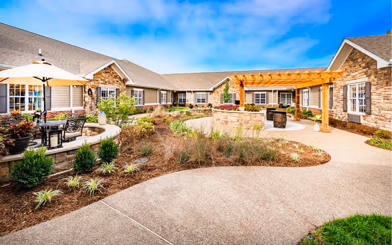 Outdoor courtyard area of a senior living facility with a paved walkway, landscaped garden beds, a wooden pergola, and seating areas including a table with an umbrella and chairs. The building exterior features stone and siding with multiple windows under a clear blue sky.