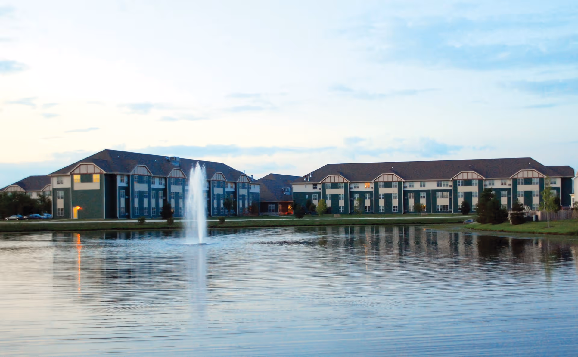 View of a senior living facility named The Timbers of Shorewood with multiple connected buildings in green and beige colors, situated behind a large pond with a water fountain in the center, under a partly cloudy sky at dusk.