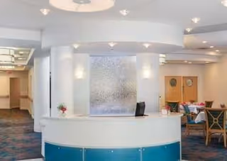 Reception area with a curved white desk featuring a blue lower panel, a textured glass panel behind the desk, and a hallway leading to other rooms. There are chairs and tables visible in the background, with soft lighting and a carpeted floor.