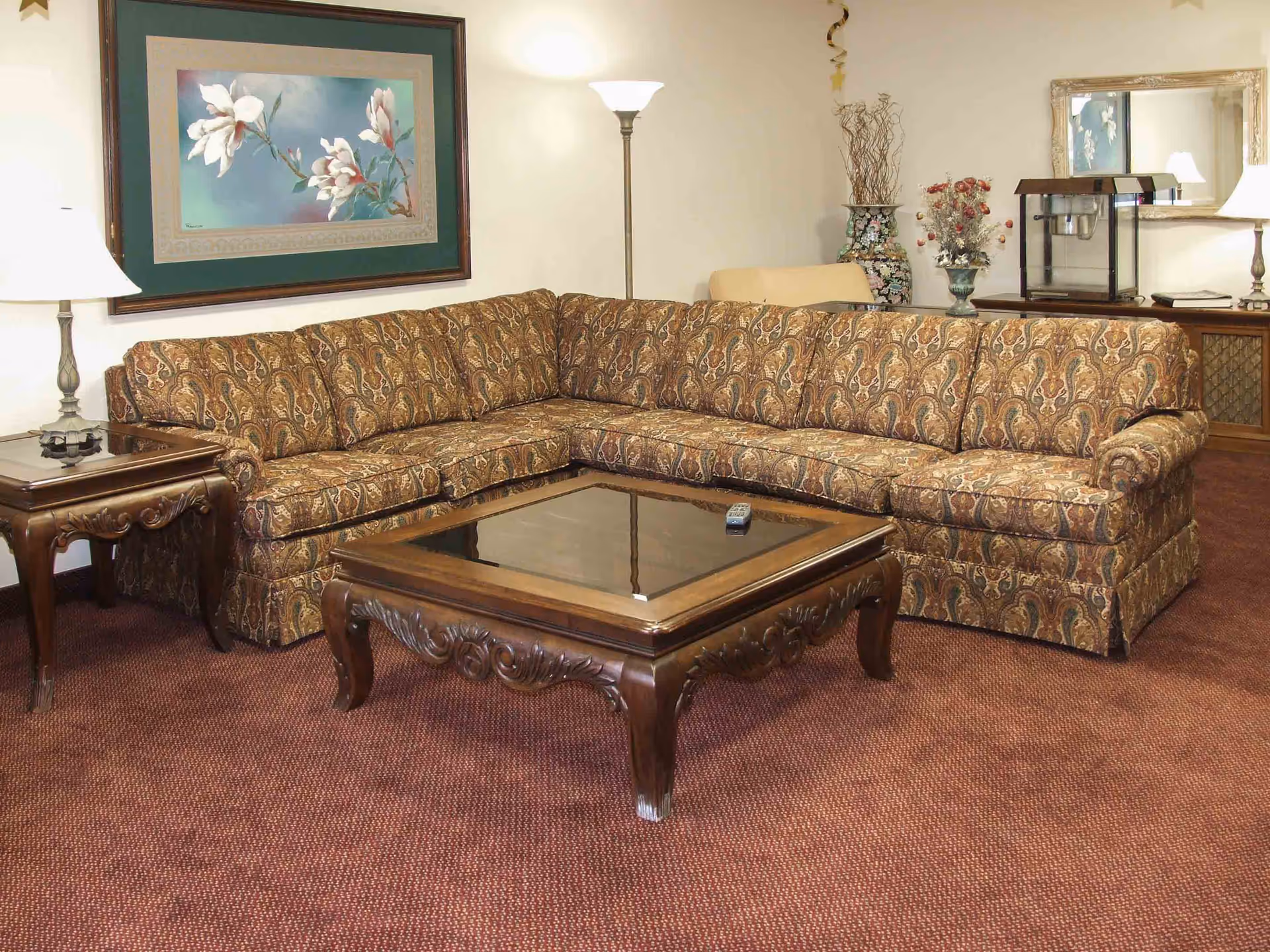 A patterned U-shaped sectional sofa and ornate wooden coffee table in a carpeted lounge with lamps, wall art, and decorative accents.