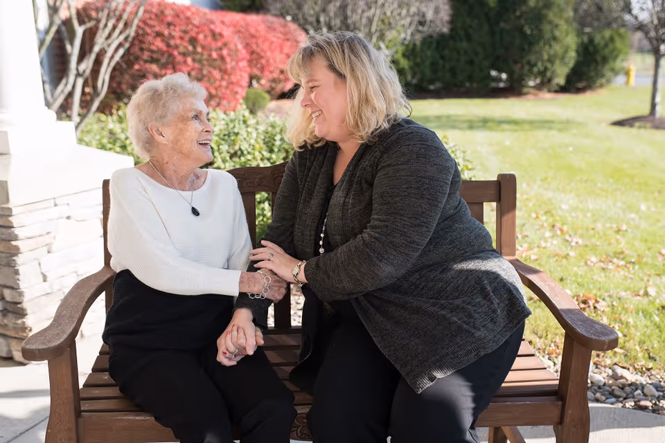 An elderly woman and a middle-aged woman sitting on a wooden bench outdoors, smiling and holding hands while engaging in a warm conversation. Behind them are green bushes and a lawn with some red foliage.