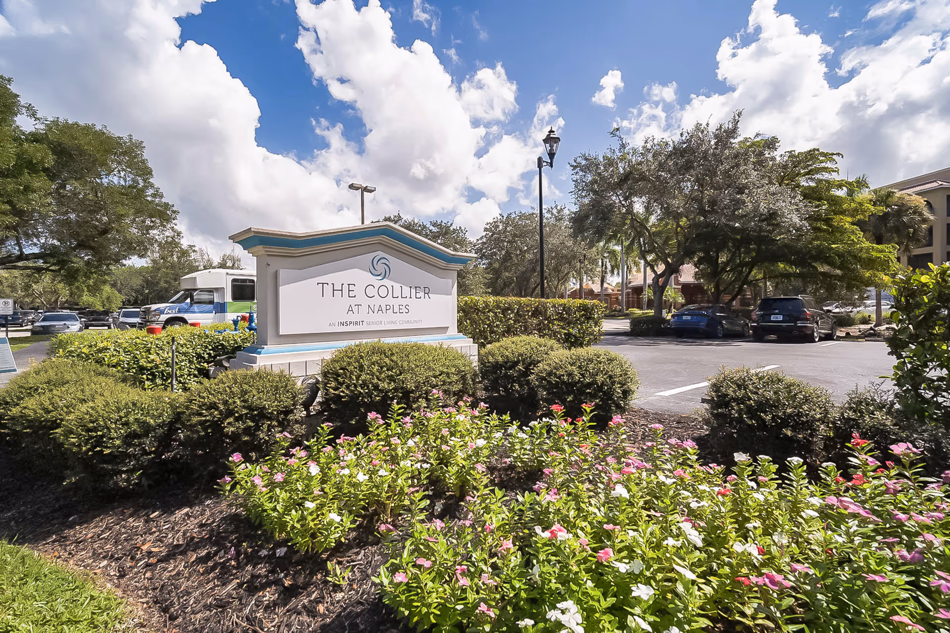Entrance sign for The Collier at Naples set among landscaped flowers and shrubs with a parking lot and trees in the background under a blue sky.