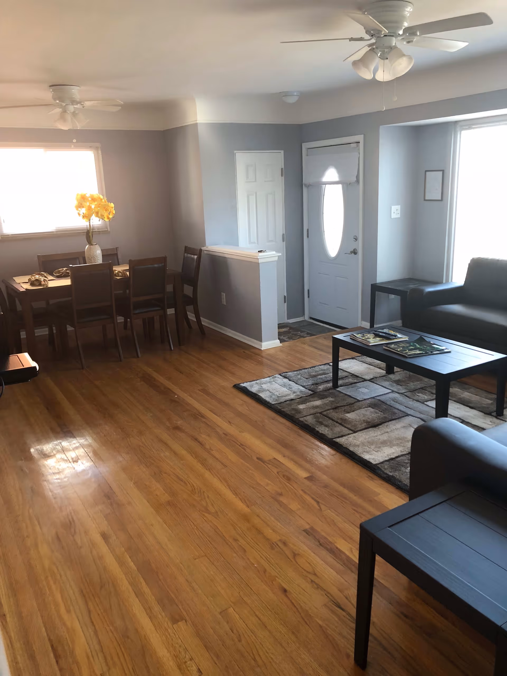 Interior view of a senior living facility room with wooden flooring, a dining table with six chairs and a vase with yellow flowers, a sitting area with two dark sofas, a coffee table with magazines, a side table, and a white front door with an oval window. The walls are painted light gray and there are ceiling fans with lights.