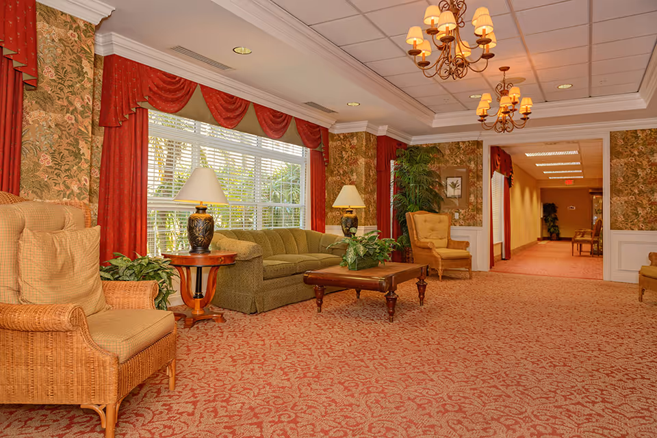 A cozy living room area with patterned red carpet and floral wallpaper. The room features a green sofa, two cushioned armchairs, two table lamps on wooden side tables, a wooden coffee table with a plant, and large windows with red curtains. Two chandeliers hang from the ceiling, and there is a hallway visible in the background.