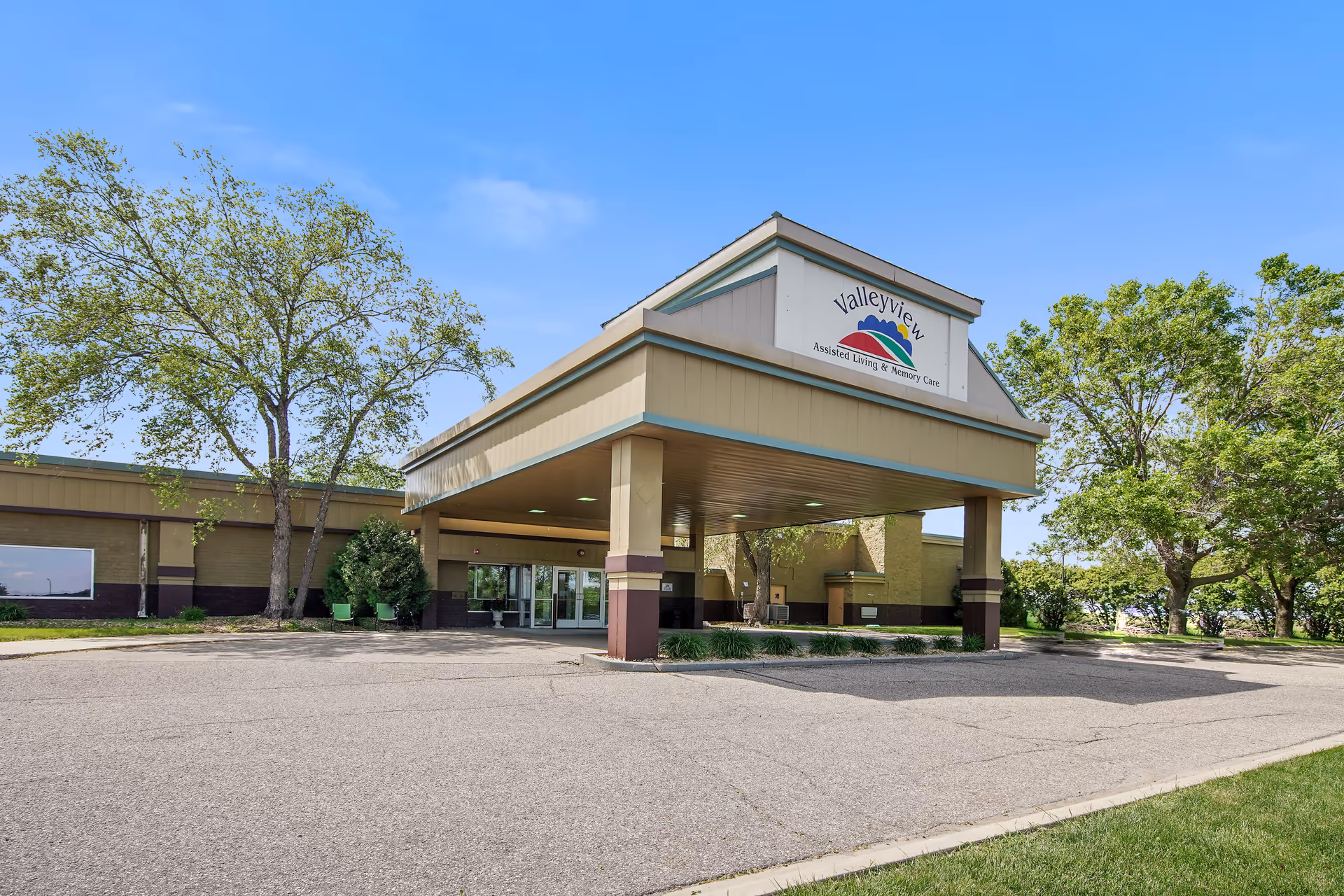 Single-story assisted living building entrance with a covered drop-off canopy labeled "Valleyview", surrounded by trees under a clear blue sky.