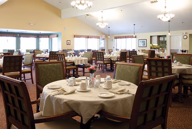 Bright, spacious dining room with round tables set for meals, wooden chairs, chandeliers, and large windows.