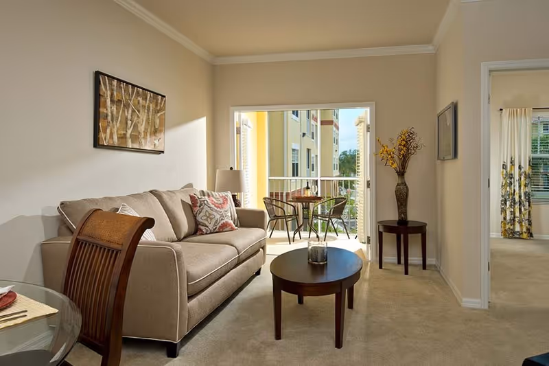 A cozy living room with a beige sofa adorned with patterned cushions, a wooden coffee table with a candle, and a side table with a vase of yellow flowers. The room opens to a balcony with a small round table and two chairs, overlooking a neighboring building. A doorway leads to a bedroom with floral curtains.
