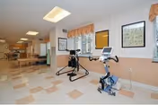 Interior view of a rehabilitation or therapy room with exercise equipment including a stationary bike and a seated elliptical machine. The room has tiled flooring, windows with valances, framed pictures on the walls, and fluorescent ceiling lights. The space appears clean and well-lit.