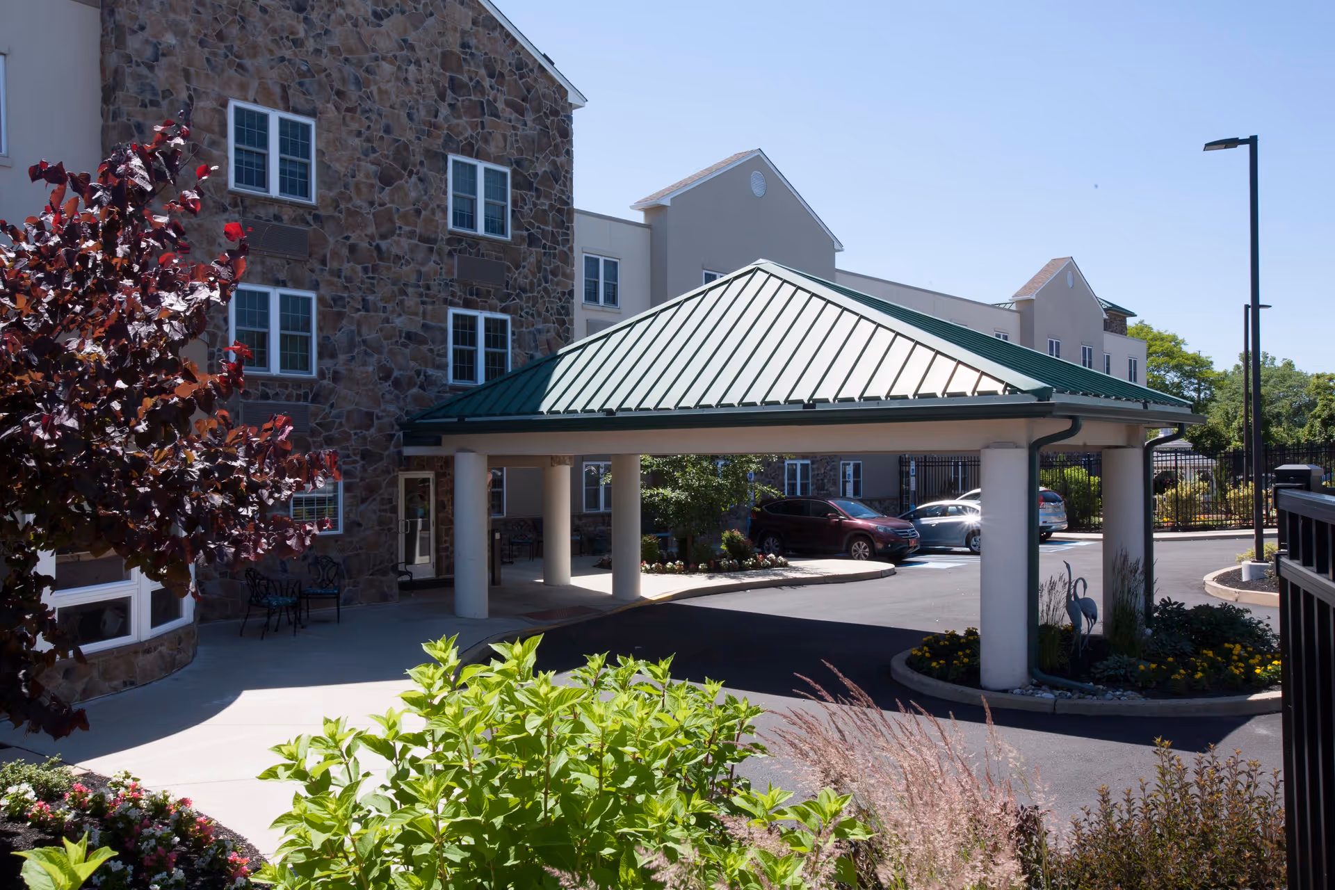 Exterior view of a senior living facility with a stone facade and a covered entrance supported by white columns. There are several windows on the building, a few parked cars, landscaped greenery, and a clear blue sky.