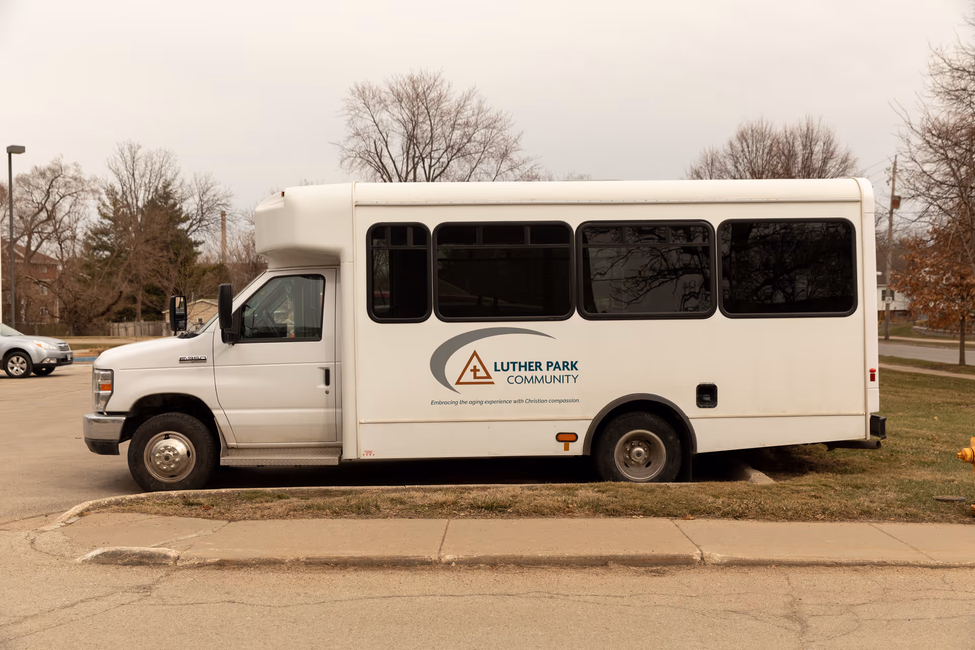 A white shuttle bus parked on a street with the logo and text 'Luther Park Community' on its side. The bus is positioned next to a grassy area and sidewalk, with leafless trees and a few other vehicles in the background under an overcast sky.