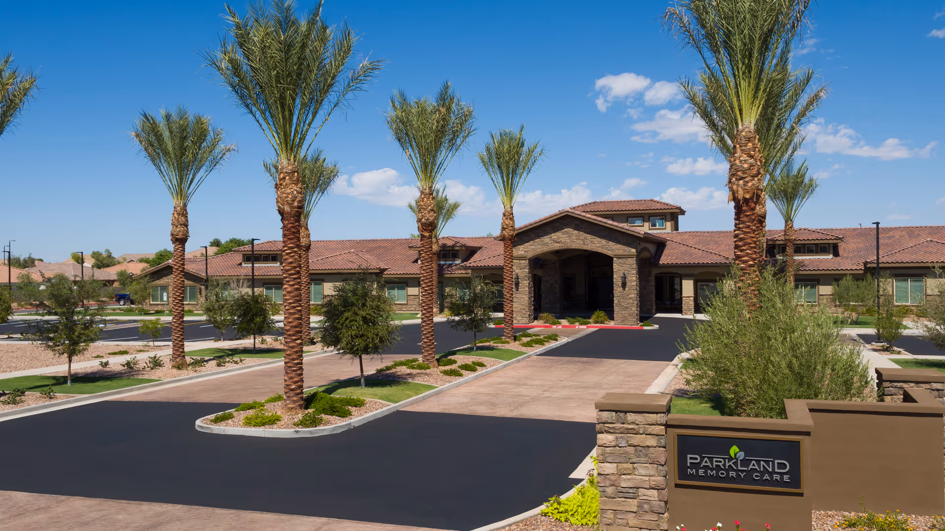 Front exterior of Parkland Memory Care building with a driveway lined by palm trees and a sign at the entrance.