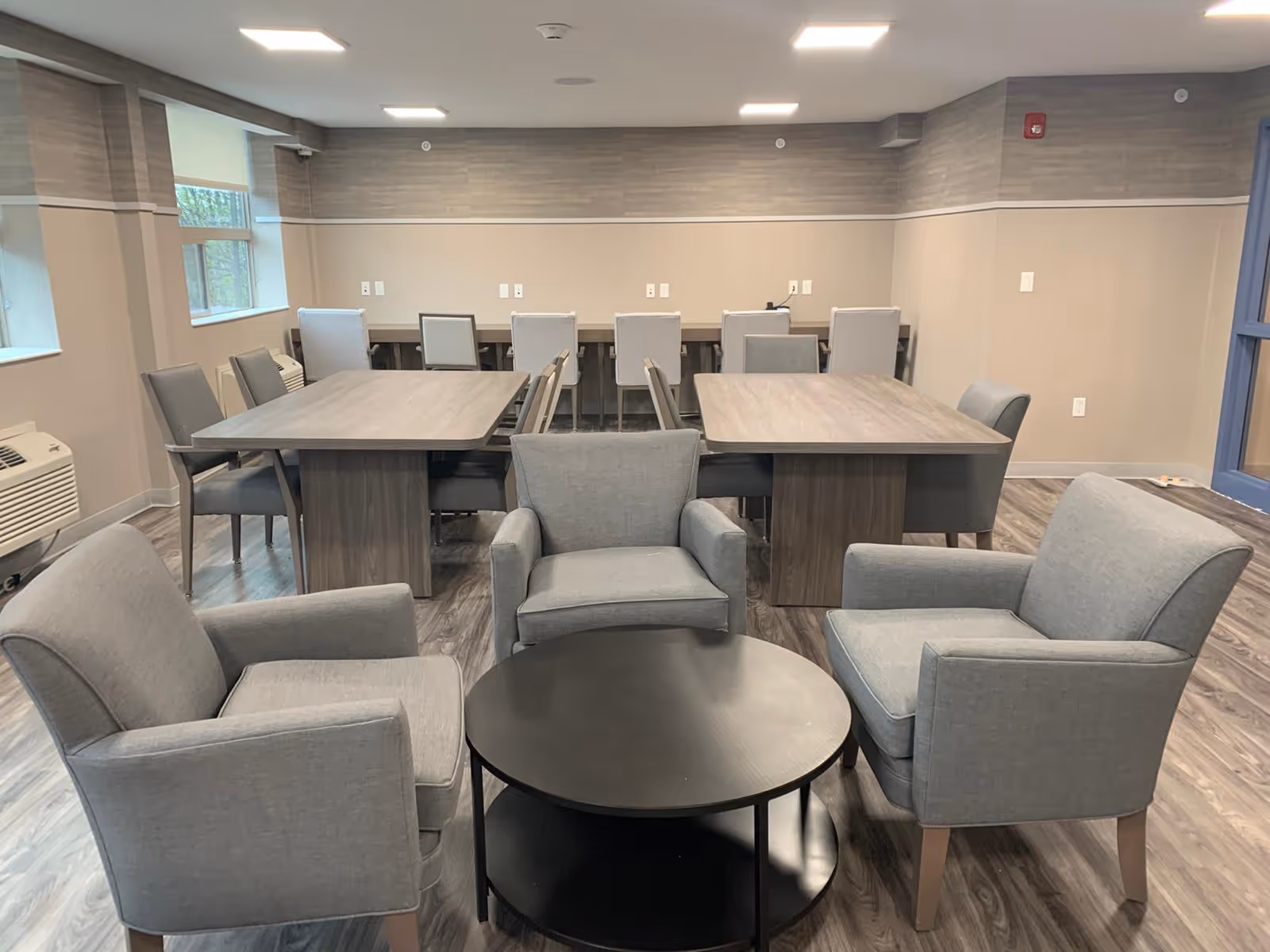 A communal room with three gray upholstered armchairs arranged around a round black coffee table in the foreground. Behind them are two rectangular wooden tables with multiple gray chairs. The room has wood-patterned flooring, beige walls with a gray accent near the ceiling, and windows on the left side.