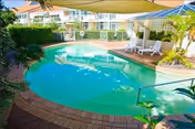 Outdoor swimming pool area with clear blue water surrounded by a brick deck. There are white lounge chairs and a large umbrella providing shade. The pool area is enclosed by a white fence and greenery, with a multi-story building visible in the background.