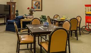 A cozy common area with multiple tables and chairs arranged for socializing or playing games. One table has a game set up on it, and there is a popcorn machine in the corner. The room also features a blue armchair, a TV, a small table with decorative items, and framed artwork on the wall.