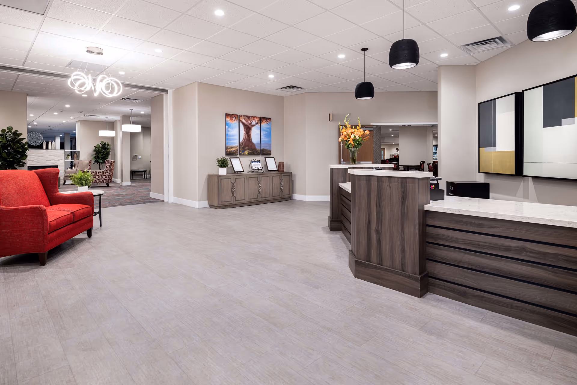 A spacious and modern reception area in a senior living facility featuring a wooden front desk with a marble countertop, three black pendant lights hanging from the ceiling, a red armchair on the left, a sideboard with framed pictures and a plant, and abstract artwork on the wall. The area is well-lit with recessed ceiling lights and has light-colored flooring.